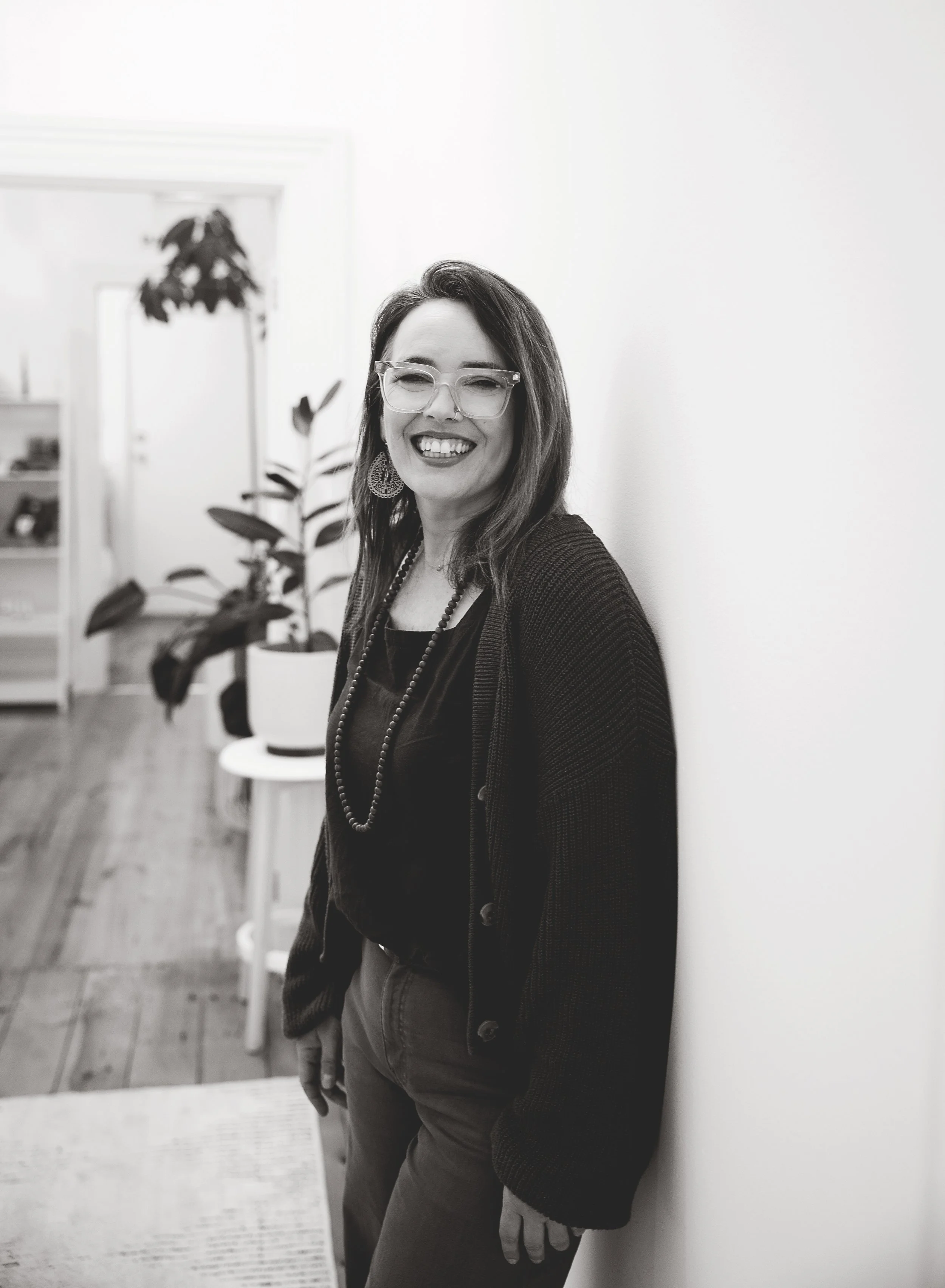 Black and white photo of a woman with glasses smiling, leaning against a plain wall in an indoor setting with a potted plant and furniture in the background.