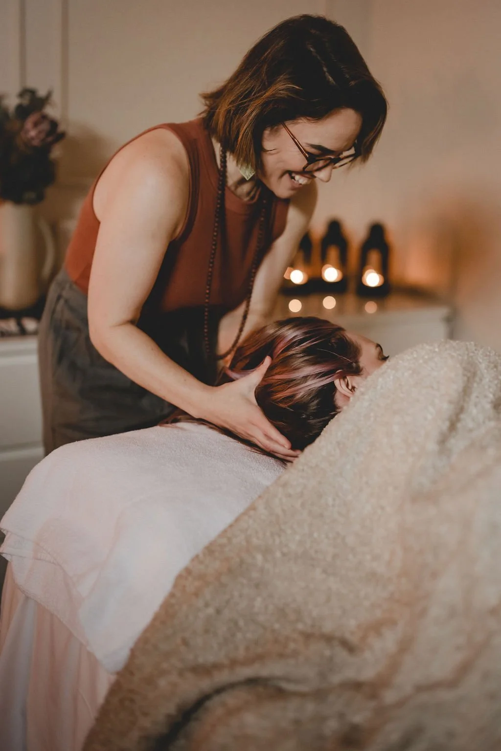 A woman giving a massage or therapy to another person lying face down on a massage table, with warm lighting and candles in the background.