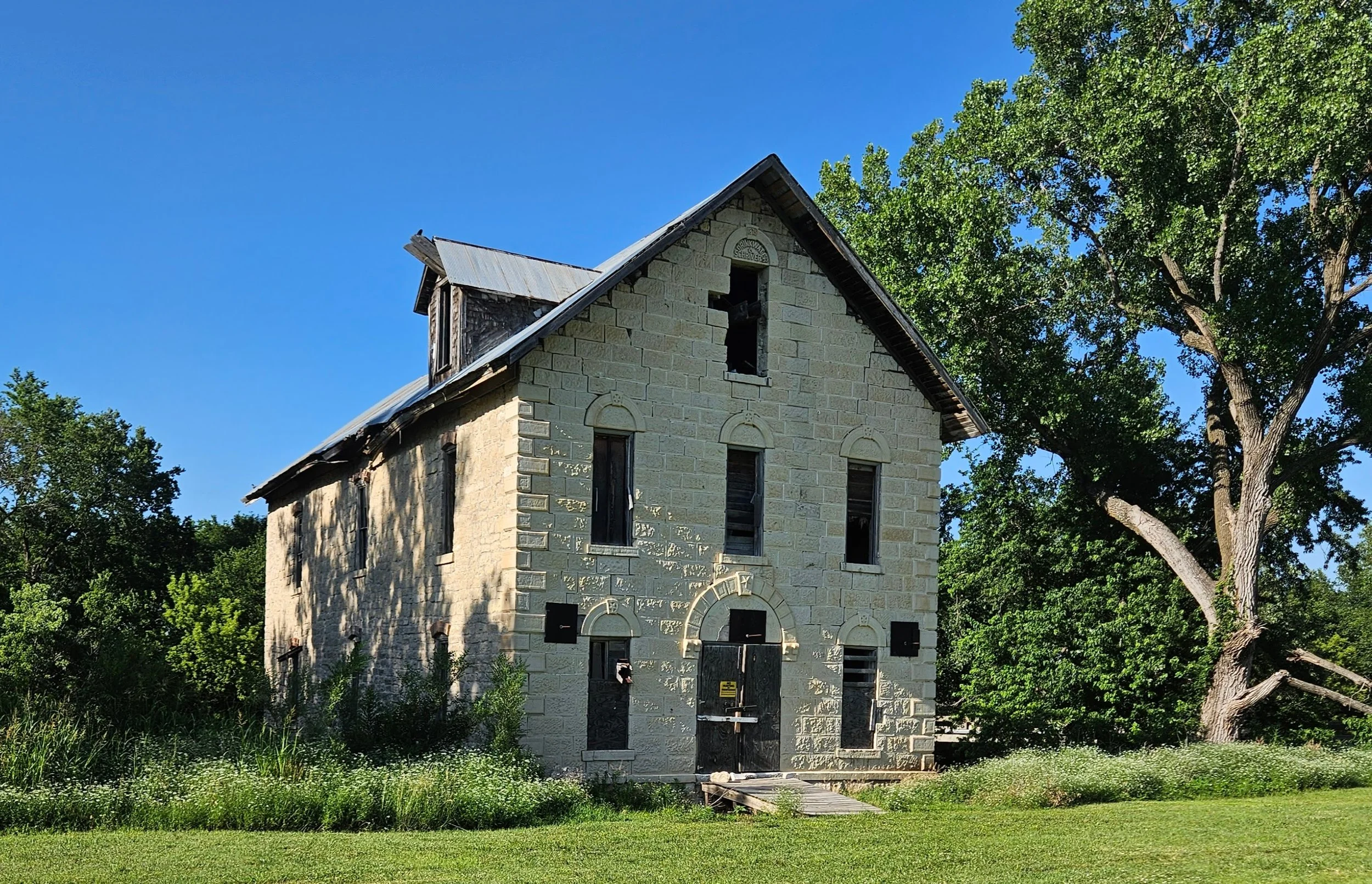 Wide view of the historic 1870s Cedar Point Mill, a native limestone structure listed on the National Register. The 3.5-story grist mill bears the name Drinkwater and Schriver on its walls, standing under a clear blue sky.