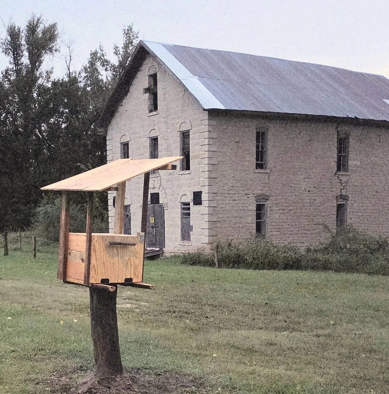 A photo of the limestone Cedar Point Mill from a distance, surrounded by grass and trees, with an empty wooden mailbox on a post in the foreground.