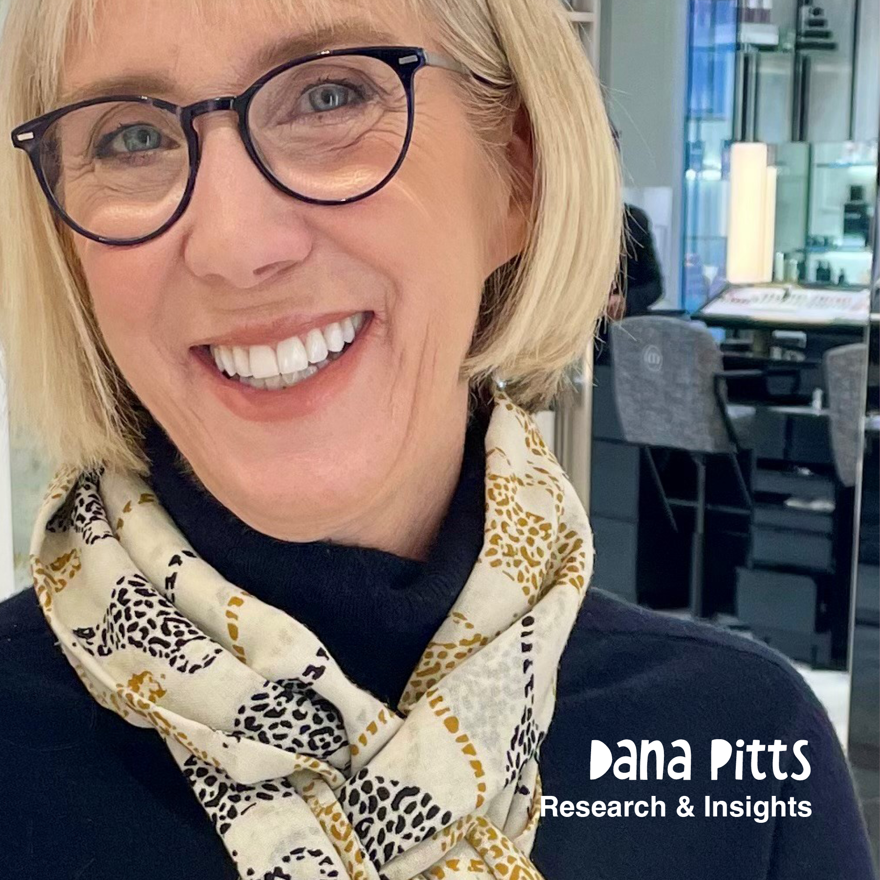A woman with short blonde hair and glasses smiling in a modern office environment, wearing a black top and a patterned scarf. The text 'Dana Pitts Research & Insights' appears in the bottom right corner.