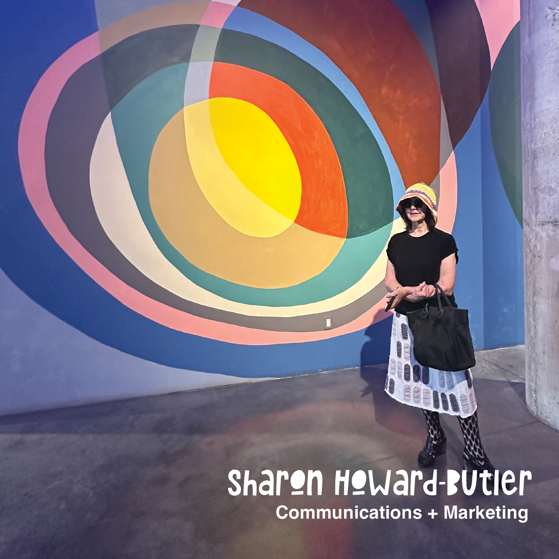A woman wearing a striped hat, sunglasses, black top, patterned skirt, and fishnet stockings standing in front of a colorful abstract mural with concentric shapes on a blue wall, holding a black bag.