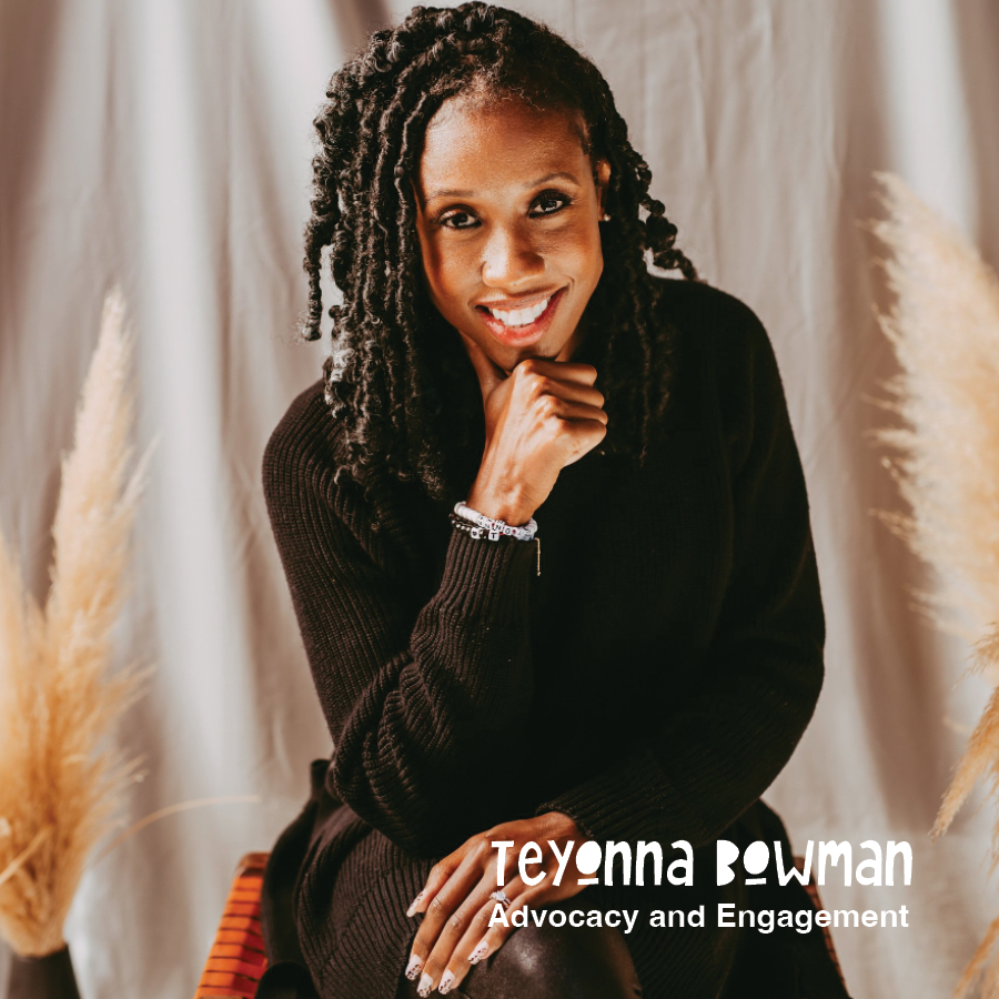 A woman with dark, curly hair and a warm smile, sitting in front of beige curtains, wearing a black sweater and some jewelry, with pampas grass in the background.