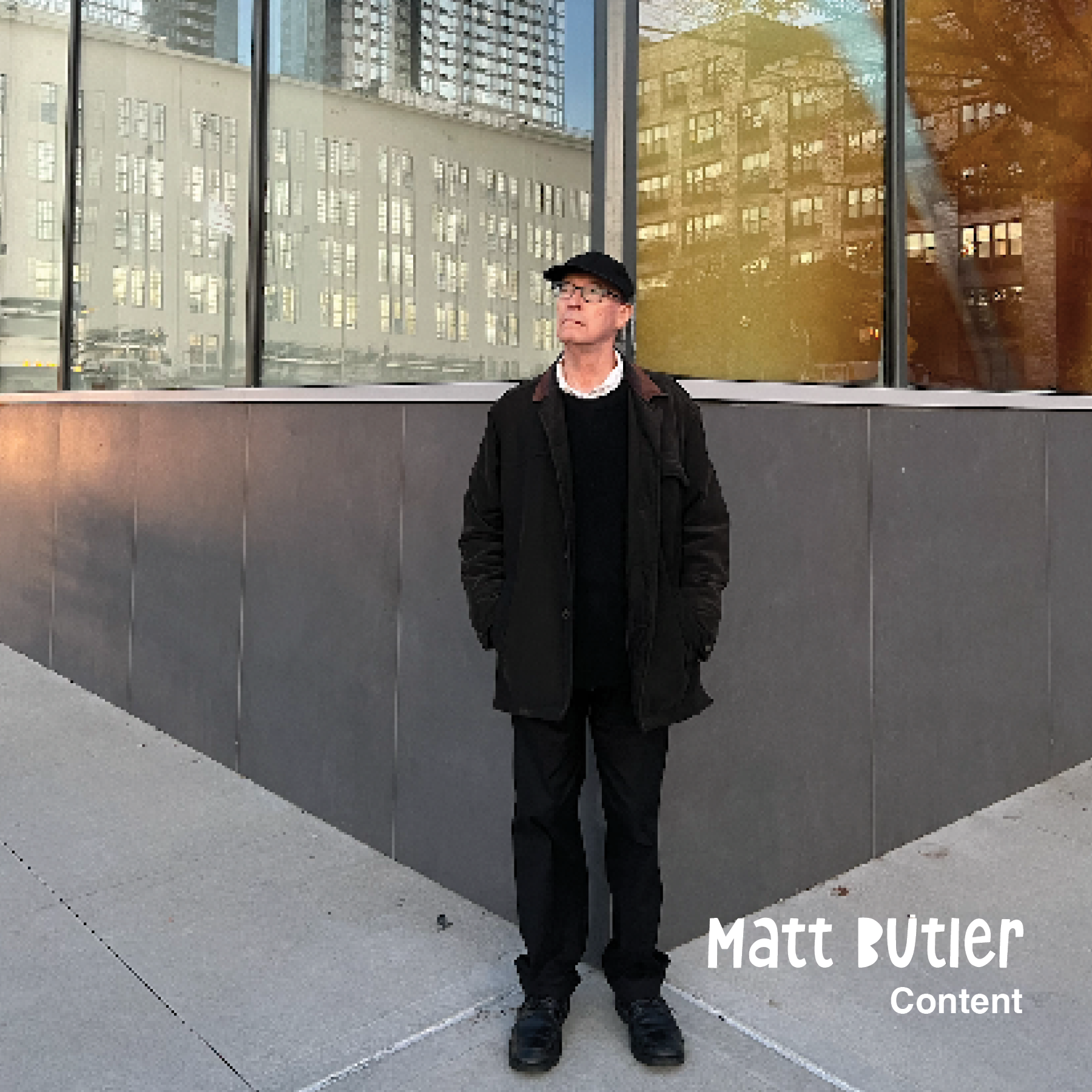 A man stands outdoors in front of a modern building with large reflective windows, wearing a black jacket, black pants, black shoes, glasses, and a black cap, with hands in his pockets, looking slightly upward.