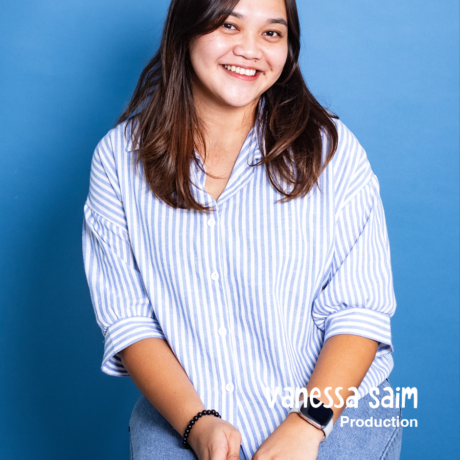Young woman with long dark hair smiling, wearing a blue and white striped shirt, standing against a blue background.