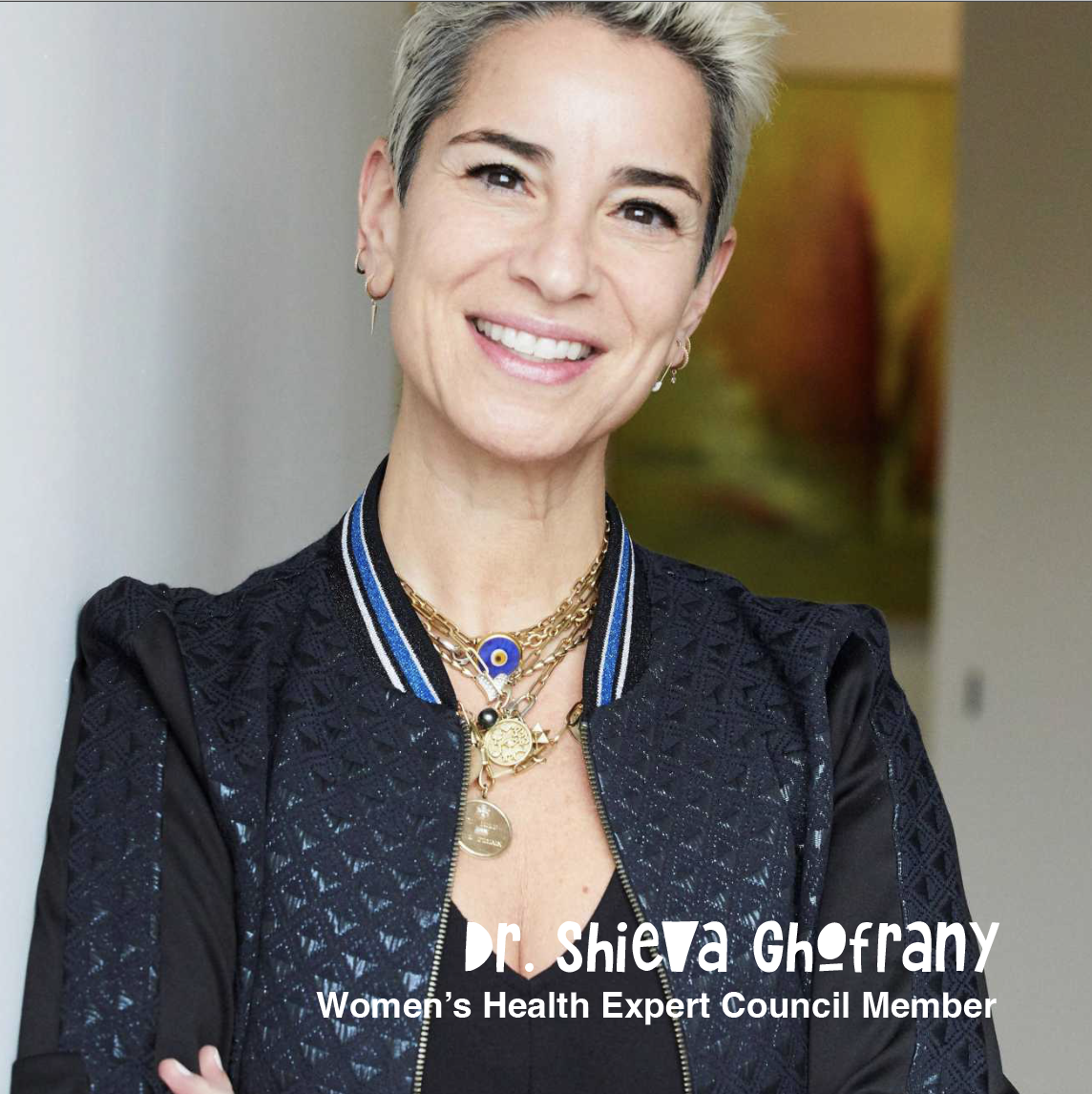 A woman with short gray hair smiling, wearing a black jacket with blue stripes on the collar and multiple gold necklaces, one with a blue and gold eye pendant. Text overlay: Dr. Shieva Ghofrany, Women's Health Expert Council Member.