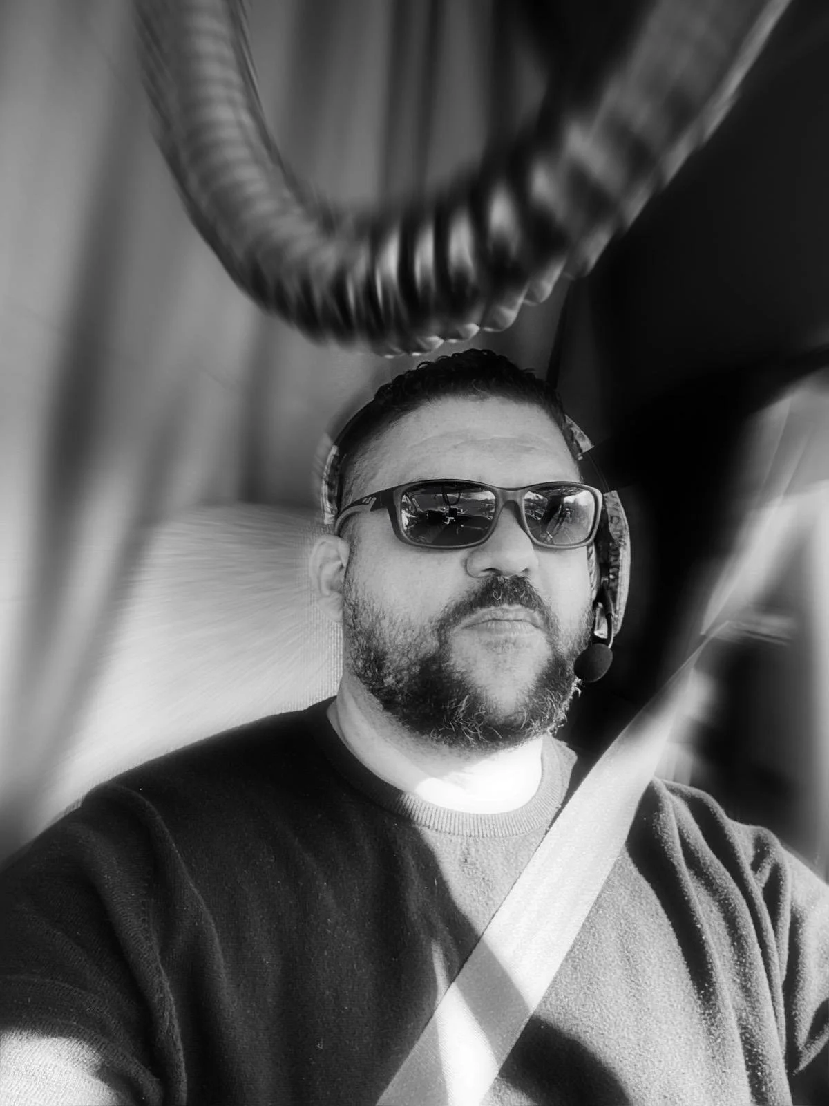 Hombre con barba, gafas de sol, auriculares con micrófono, conduciendo un vehículo, imagen en blanco y negro.