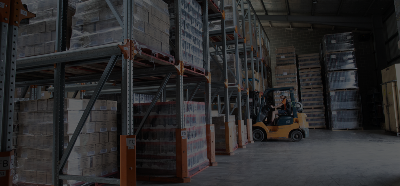 A warehouse with high metal shelving filled with pallets of boxes, some wrapped in plastic, and a forklift operator moving pallets.