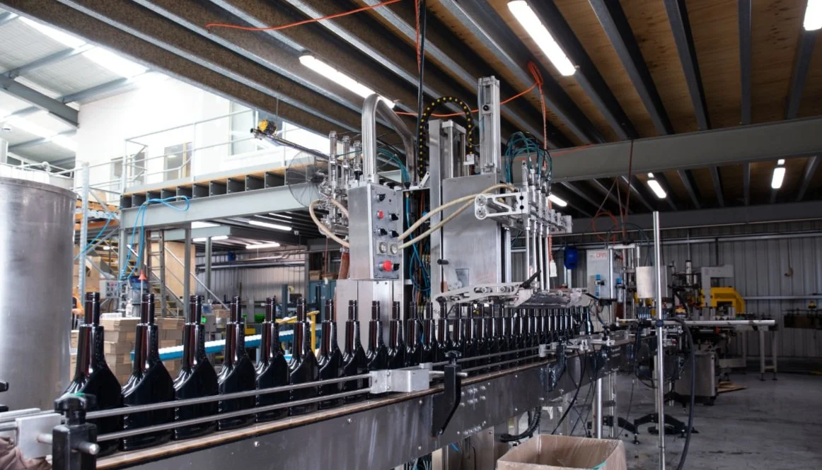 Industrial bottle filling machine in a warehouse setting. Bottles are lined up on a conveyor, ready to be filled.
