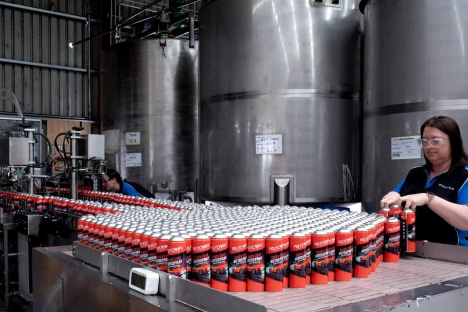 Workers in a factory handling red spray paint cans on a production line with large stainless steel tanks in the background.