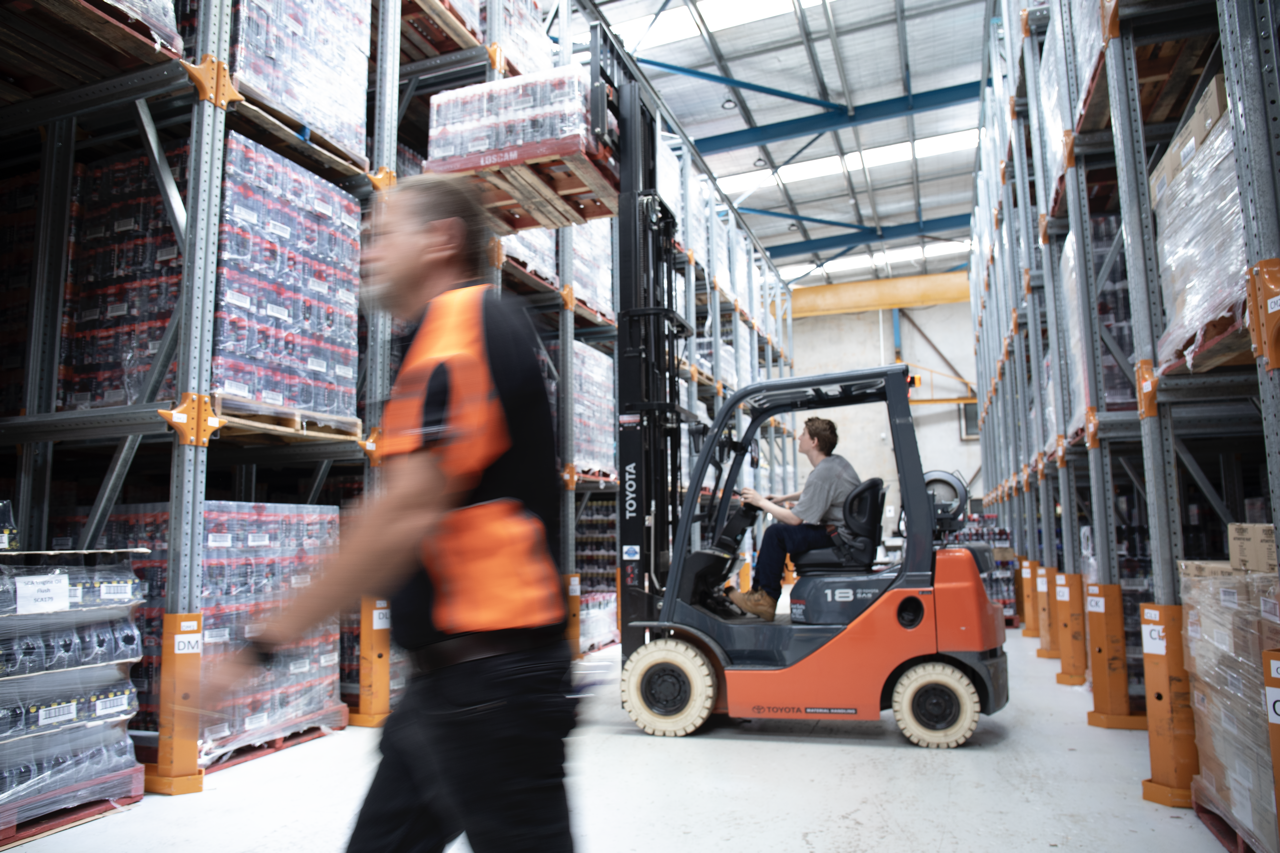 A warehouse with high shelves stocked with pallets of goods, a person in a gray shirt operating a forklift, and another person walking past in an orange and black uniform.