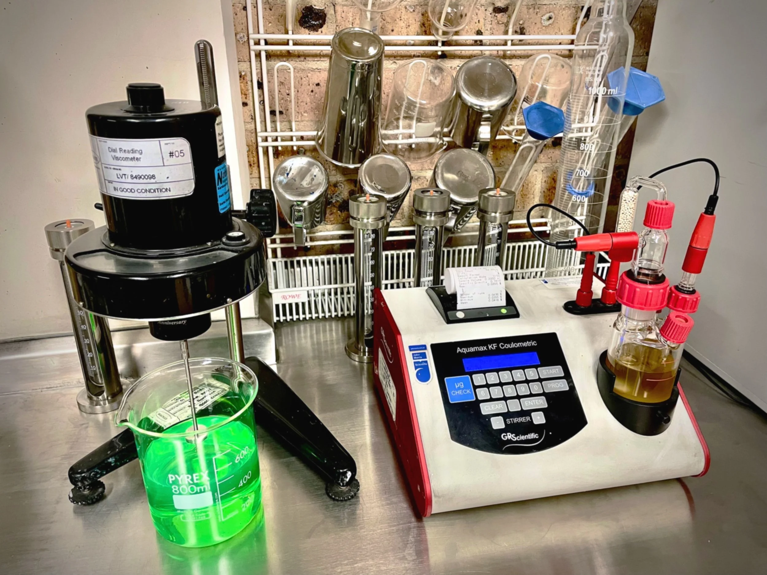 Laboratory setup with a beaker of green liquid, a viscometer, test tubes, measuring cups, and scientific equipment for liquid analysis.