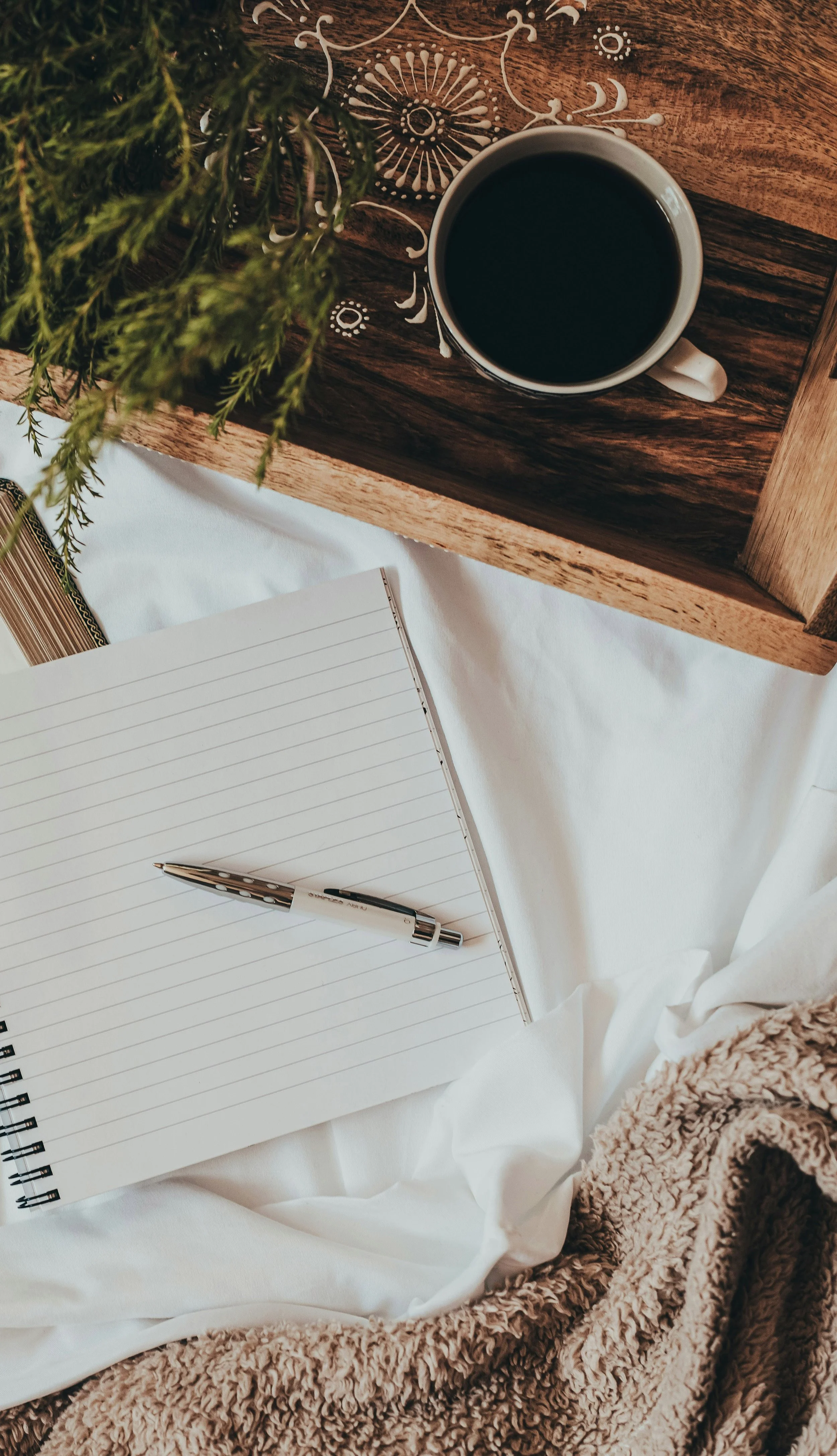 A mug of black coffee on a wooden tray with a decorative pattern, a lined notebook with a pen, a plush blanket.