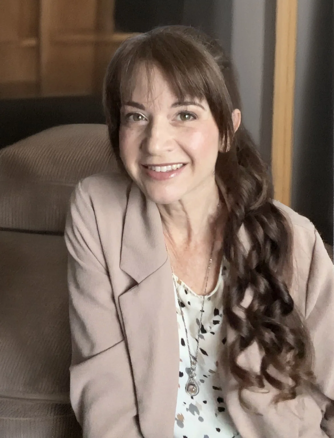 Smiling middle-aged woman with long, wavy brown hair, wearing a light beige blazer over a patterned blouse, sitting on a sofa in a warmly lit room.