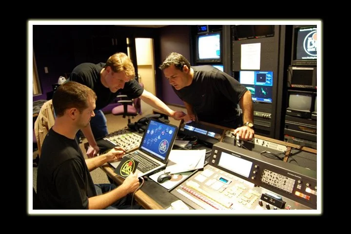 Three men working in a television or video production studio with monitors, a mixing console, and various electronic equipment.