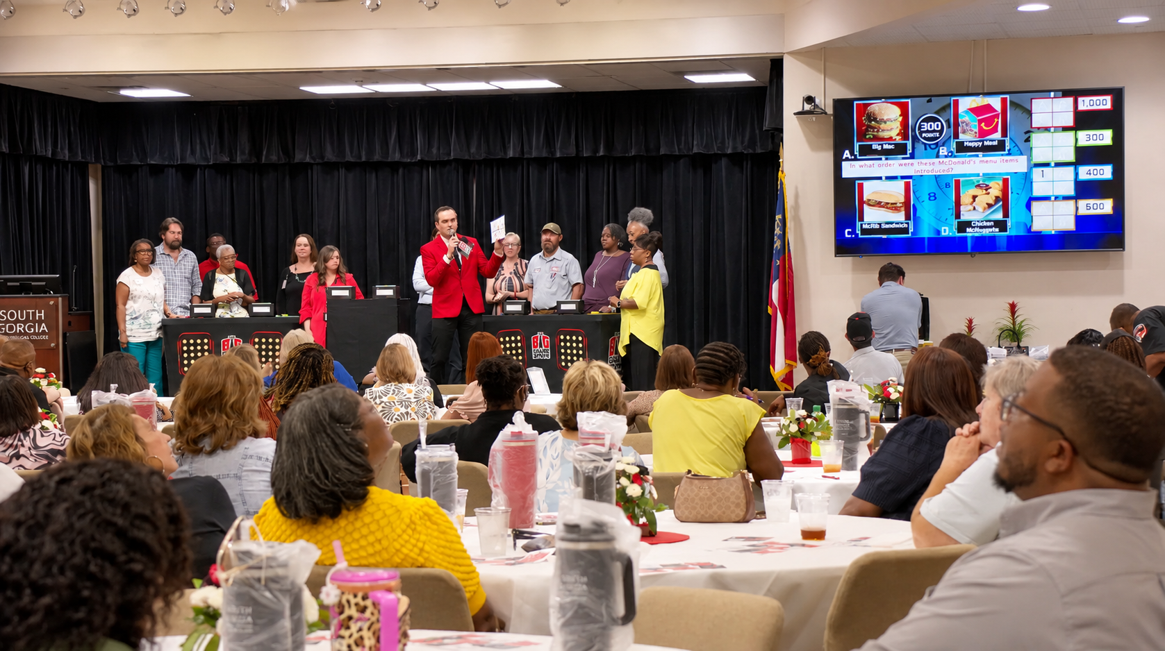 A room packed with people listening to a game show take place on stage. The stage has a panel with multiple contestants and a host in a red blazer. A large screen displays menu items from McDonald's and a quiz question.