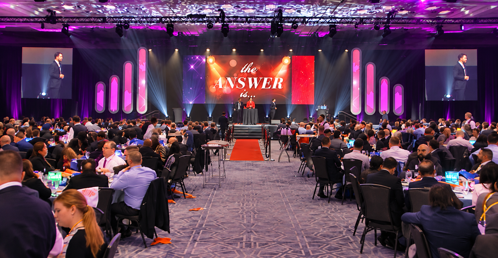 A large conference hall filled with attendees seated at round tables, a stage with a presentation screen displaying 'the answer is...', and two large screens showing a speaker in a suit.