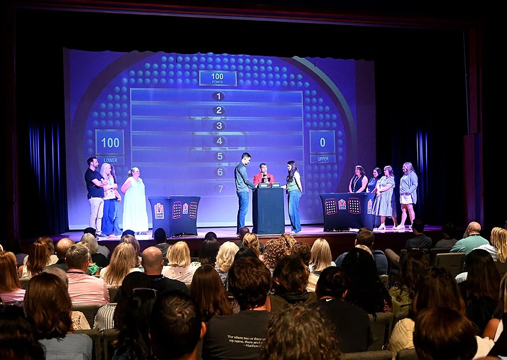 A game show set with contestants and host, large screen displaying game options, audience watching from front of the stage.