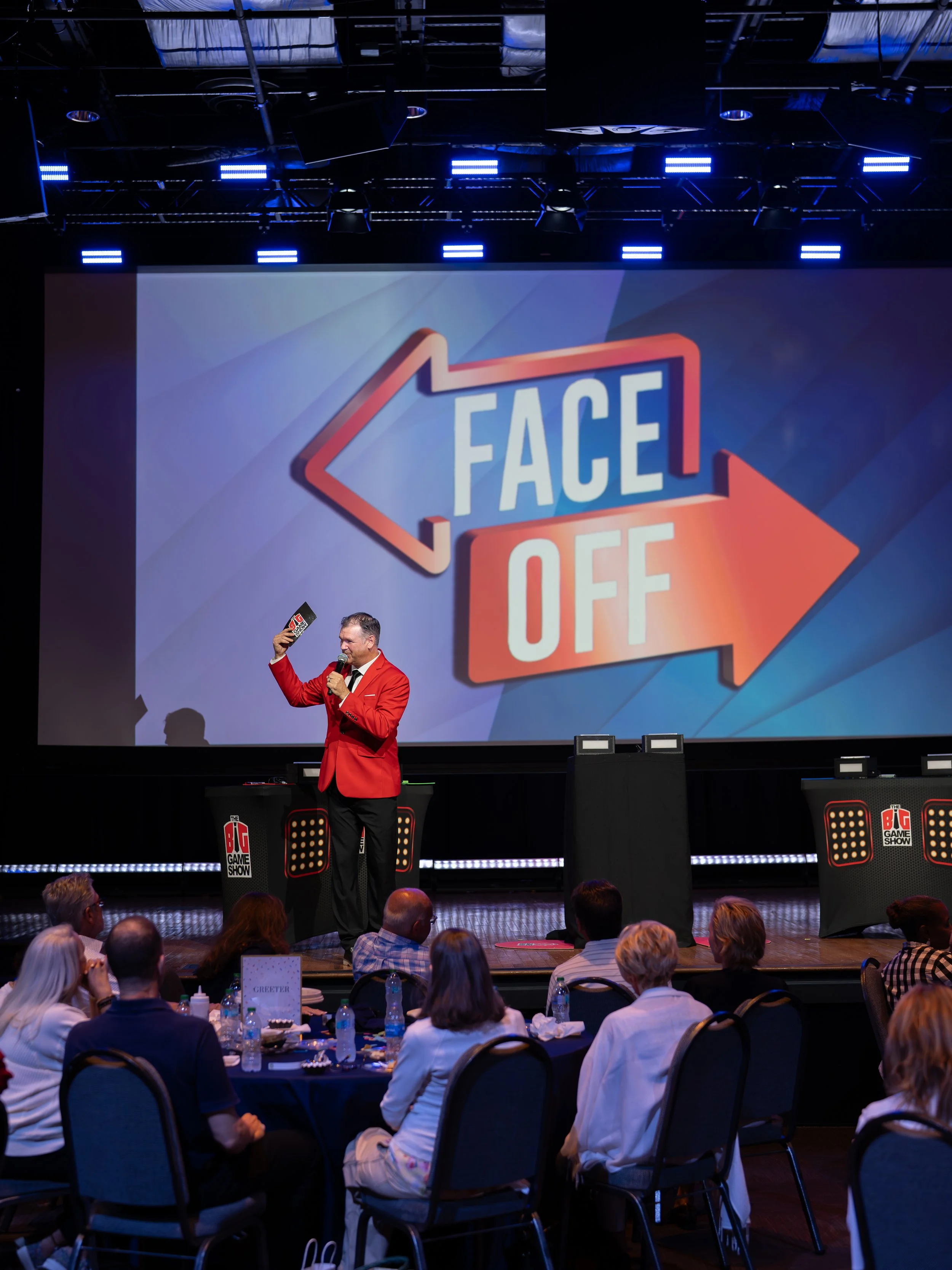 A man wearing a red blazer speaks into a microphone on stage at a conference or event, with a large screen displaying the words 'FACE OFF' behind him. Audience members are seated at tables facing the stage.