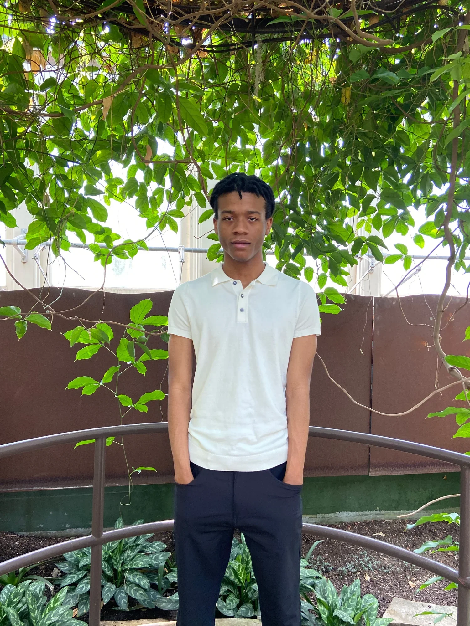 A black male posed with his hands in his pant pockets in a white polo and black dress pants, surrounded by greenery from an overarching tree.
