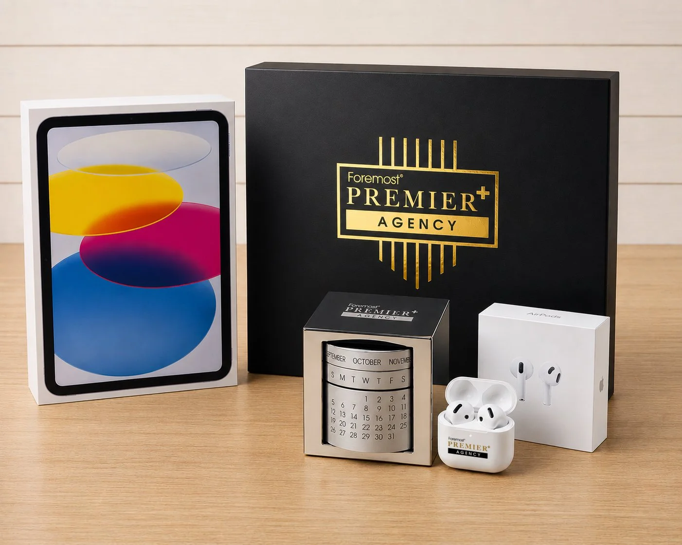 A product image of an iPad, custom-made black box with the Foremost Premier+ Agency logo on it in gold foil. A rolling desktop calender made of stainless steel, and a open pair of airpods with a airpods box propped up behind them.
