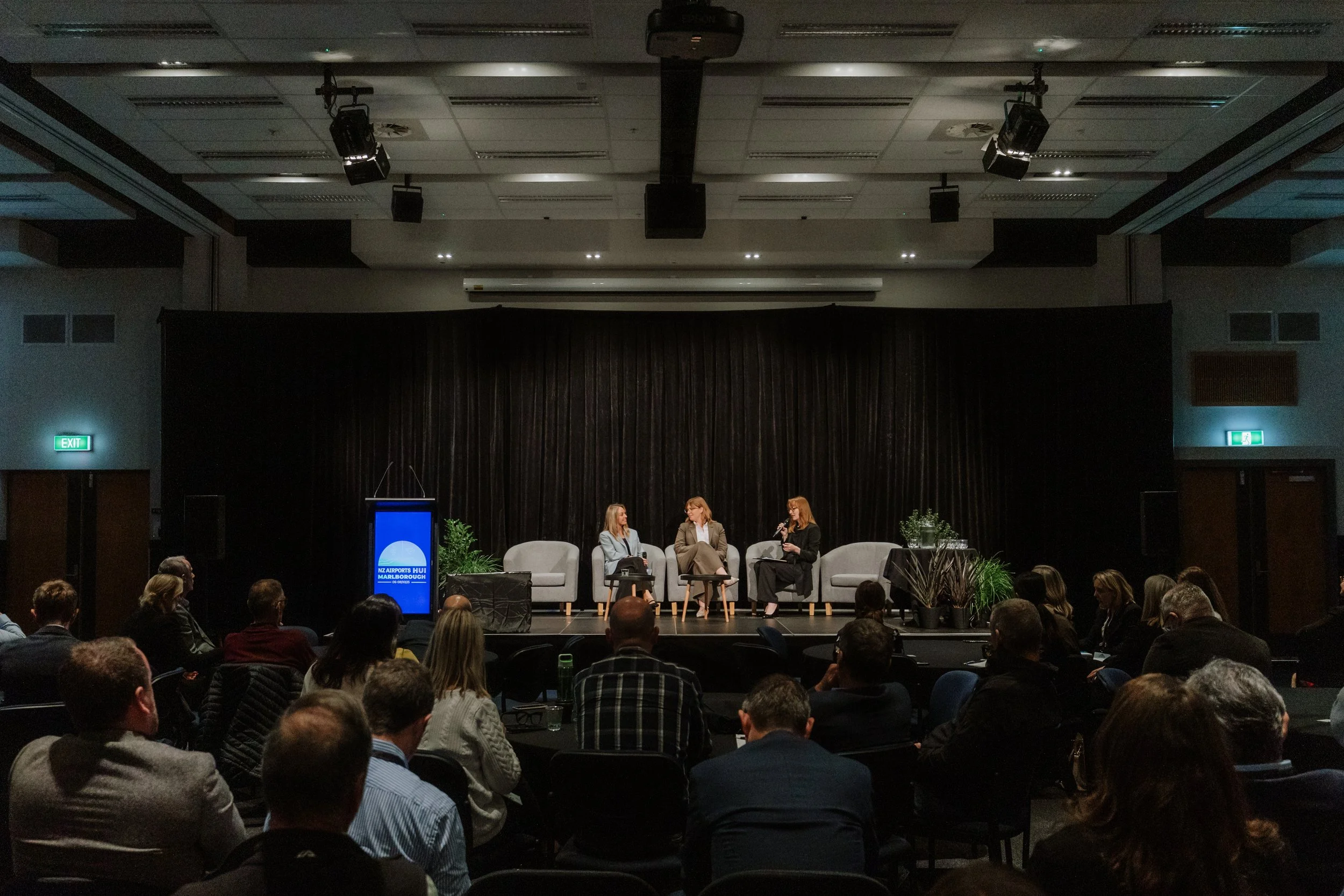 A panel discussion on a stage with four women sitting on chairs in front of a black curtain. There are plants on the stage, and a large audience is seated watching. The room has a high ceiling with stage lights and speakers. There is a blue sign with white text that reads 'NZ Airports HUI MARLBOROUGH'.
