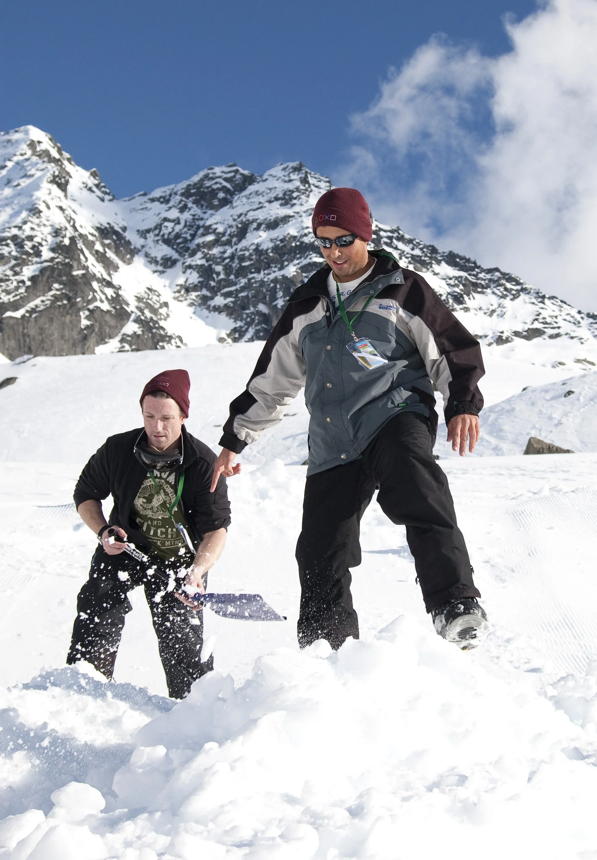 Two men with beanies and sunglasses stand in snowy mountains, one is shoveling snow, and the other is holding a snow shovel, all surrounded by snow and mountain peaks.