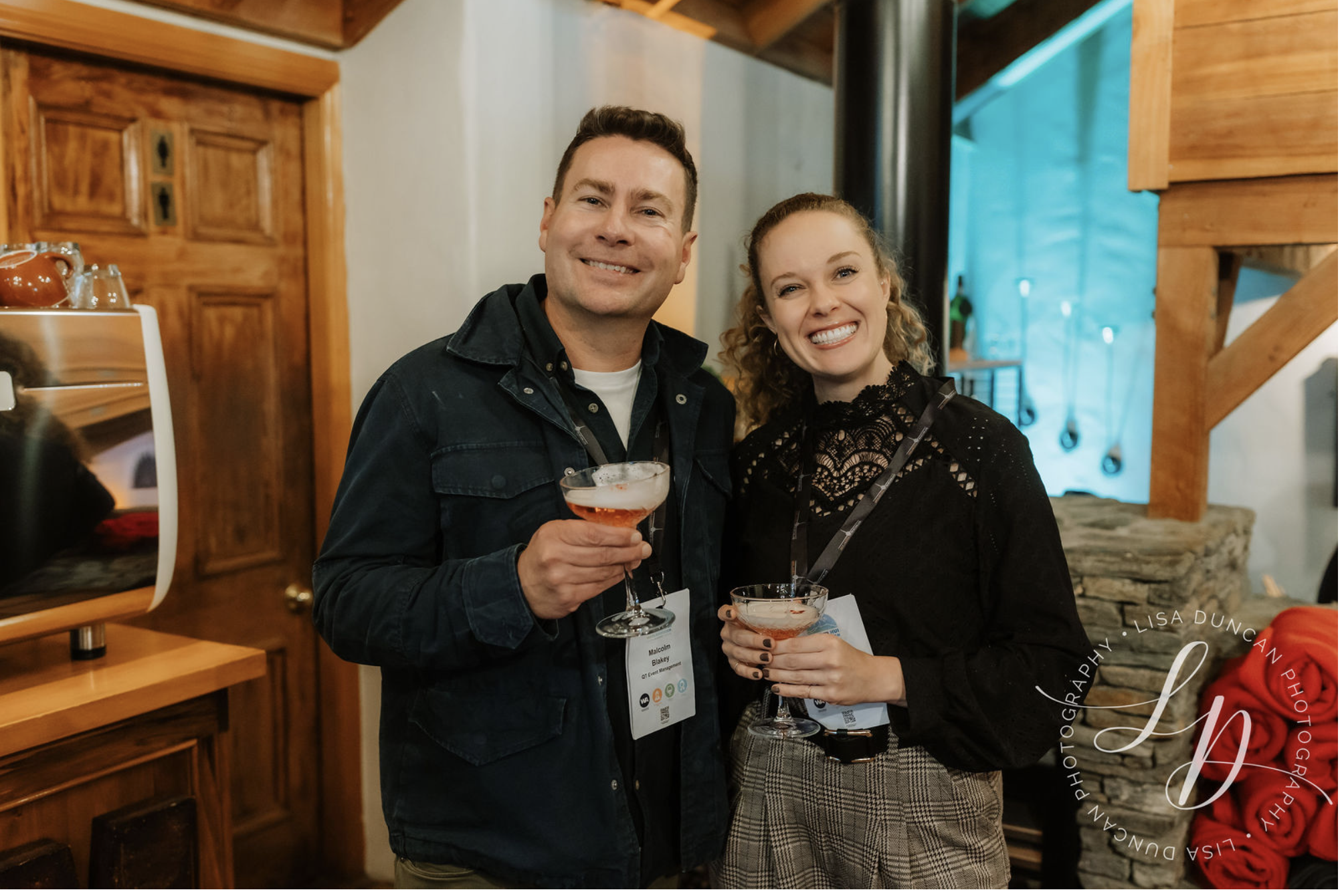 Two people smiling indoors, holding cocktail glasses with pink drinks, in a cozy wooden interior. The man is wearing a black jacket and the woman is wearing a black lace top and plaid pants.