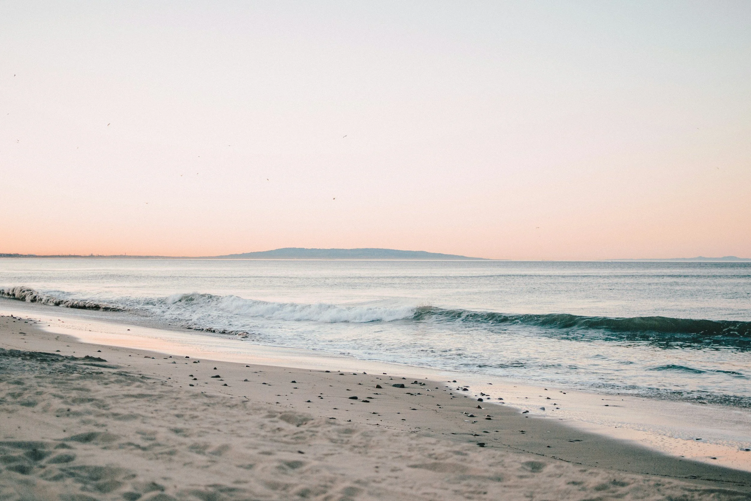 A peaceful beach scene with gentle waves, sandy shore, and a distant landmass under a soft, pastel sky.