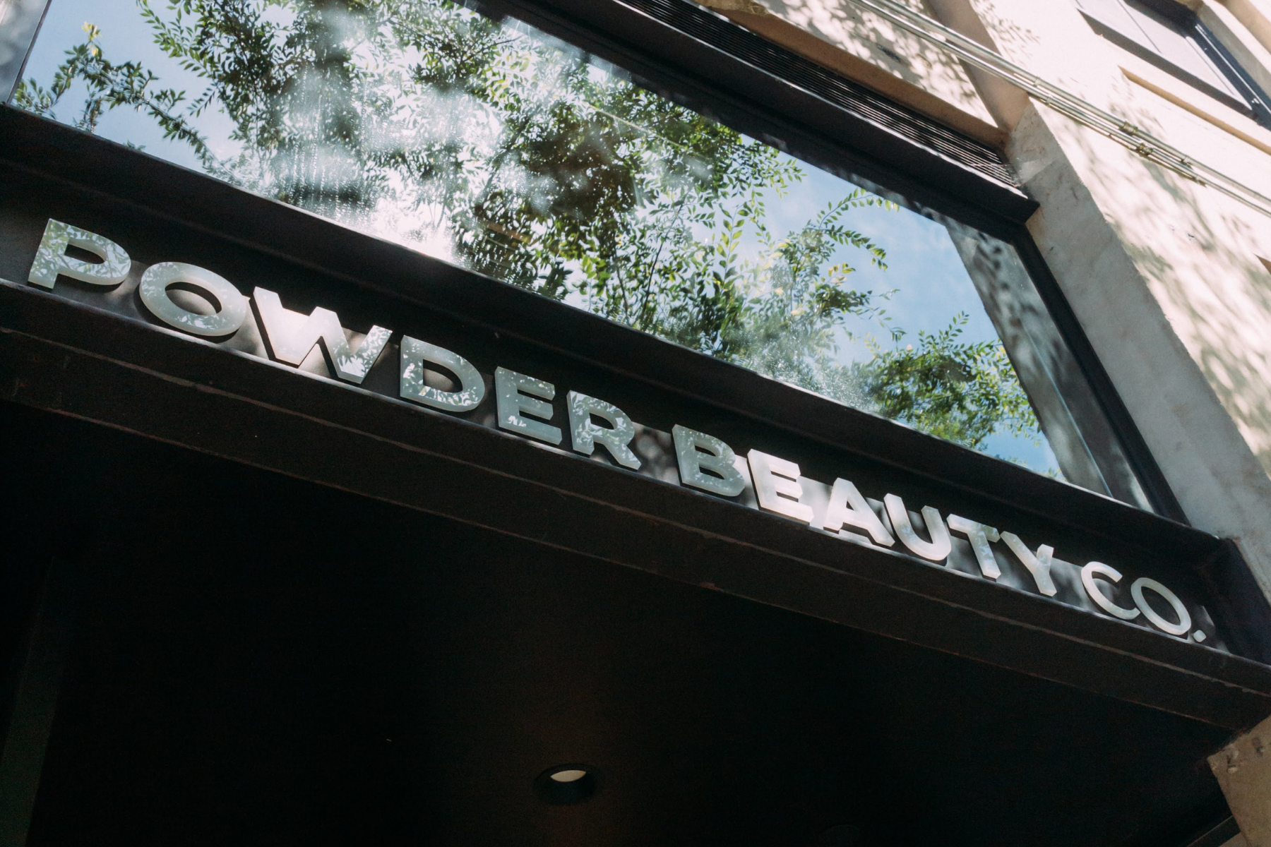 Photo of a storefront sign reading 'POWDERBEAUTY CO.' with a large window reflecting trees and blue sky above.