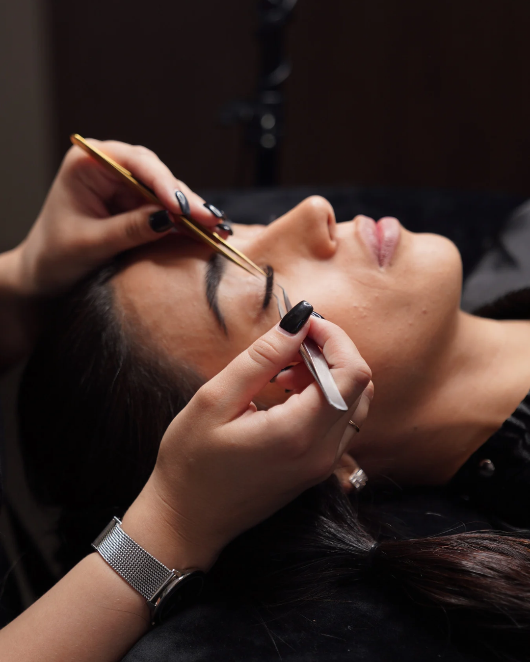A woman is lying down while receiving an eyebrow threading or shaping treatment from an aesthetician holding tools.