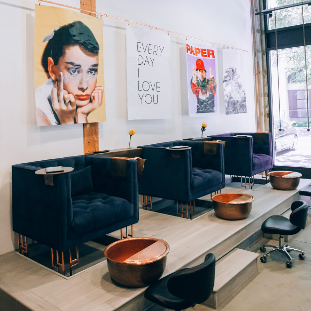 Coffee shop interior with four blue armchairs on a raised wooden platform, small tables with copper bowls, art posters on the wall including a portrait, a handwritten note that says 'EVERY DAY I LOVE YOU', and a large window showing a parked car outside.