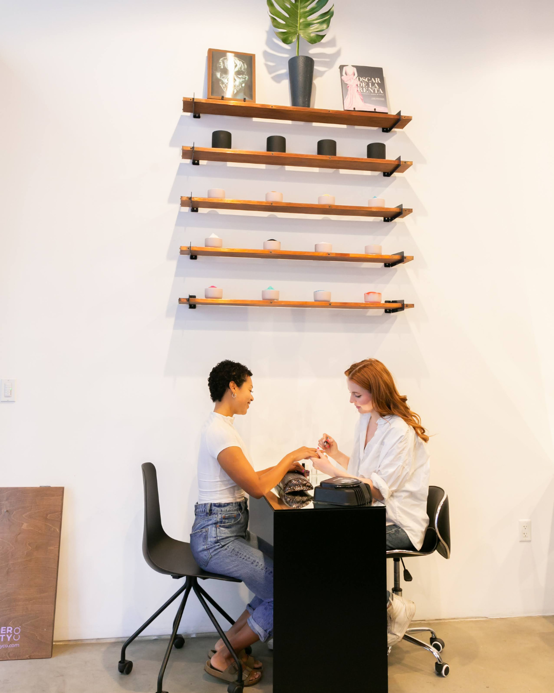 Two women sitting at a nail salon nail station, one getting a manicure. Behind them are four wooden shelves with candles, framed artwork, a plant, and a magazine on the wall.