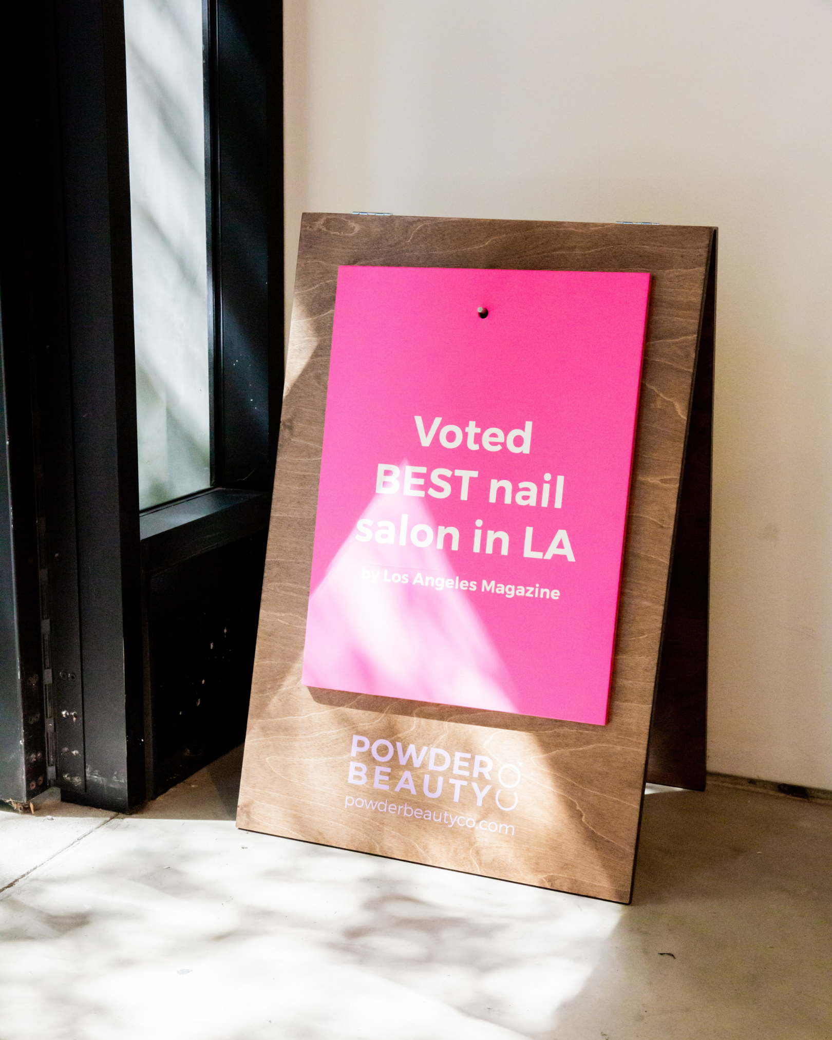 Pink signboard leaning against a brown wood stand, promoting a beauty salon in LA, with text in white reading 'Voted Best nail salon in LA by Los Angeles Magazine', located near a black window or door frame.