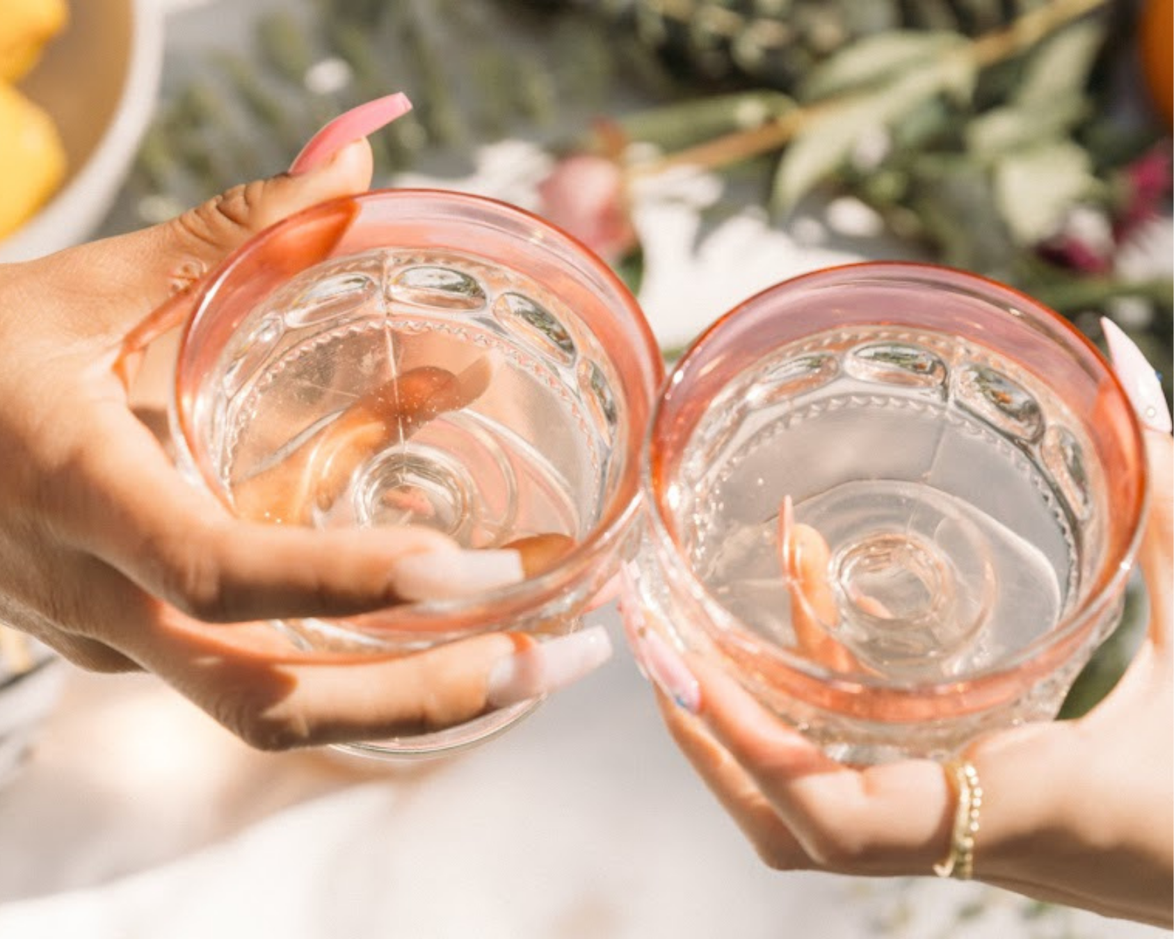 Two people holding transparent glasses with pink rims, filled with clear water, raising them in a toast outdoors. There are blurred flowers and greenery in the background.