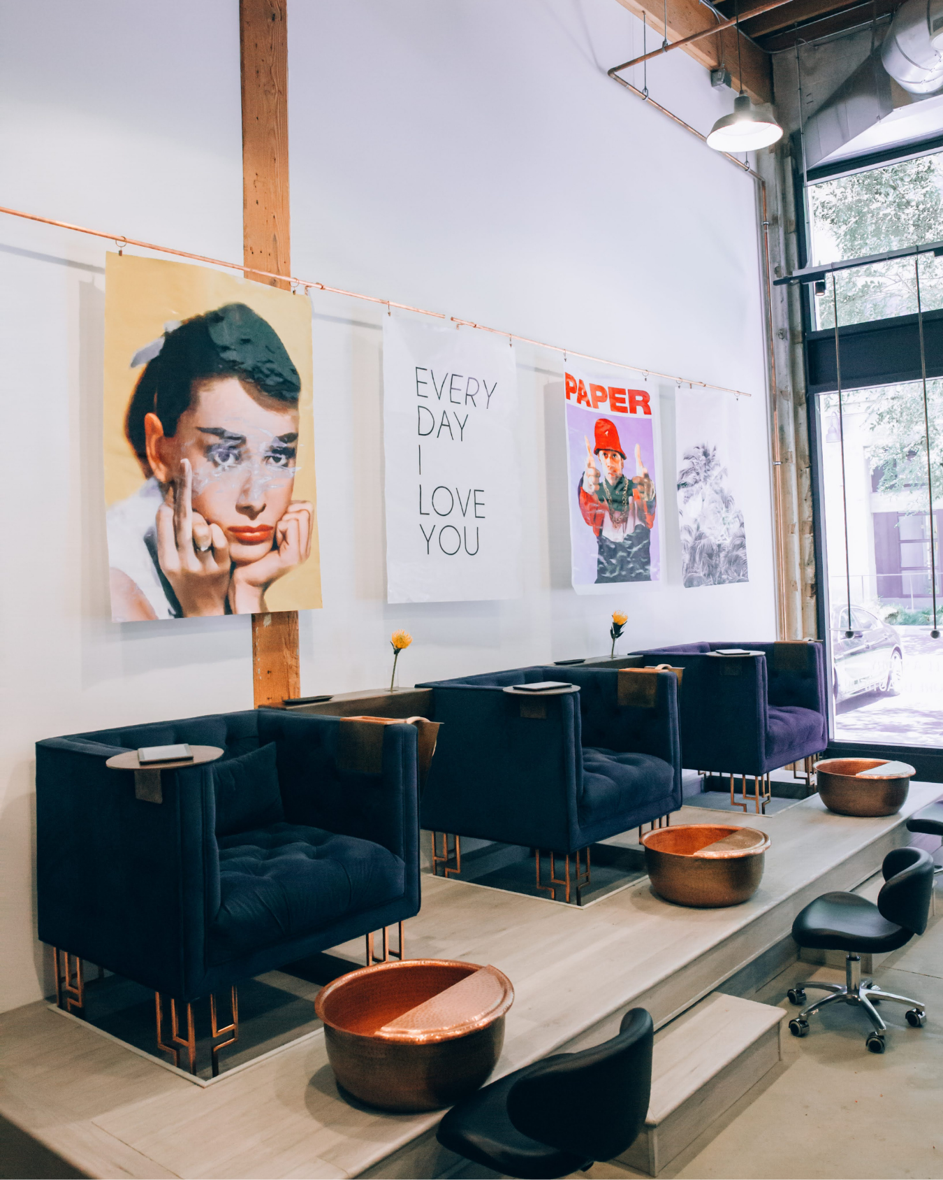 A modern salon or waiting area with black cushioned chairs, small round tables, and wall art including a vintage-style portrait, a sign that reads 'Every Day I Love You,' and colorful posters. There are large windows and wooden accents.