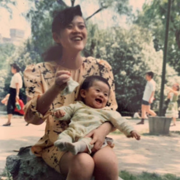 A woman sitting outdoors on a rock, holding a smiling baby on her lap, both appearing happy. The background shows trees and a few people walking.