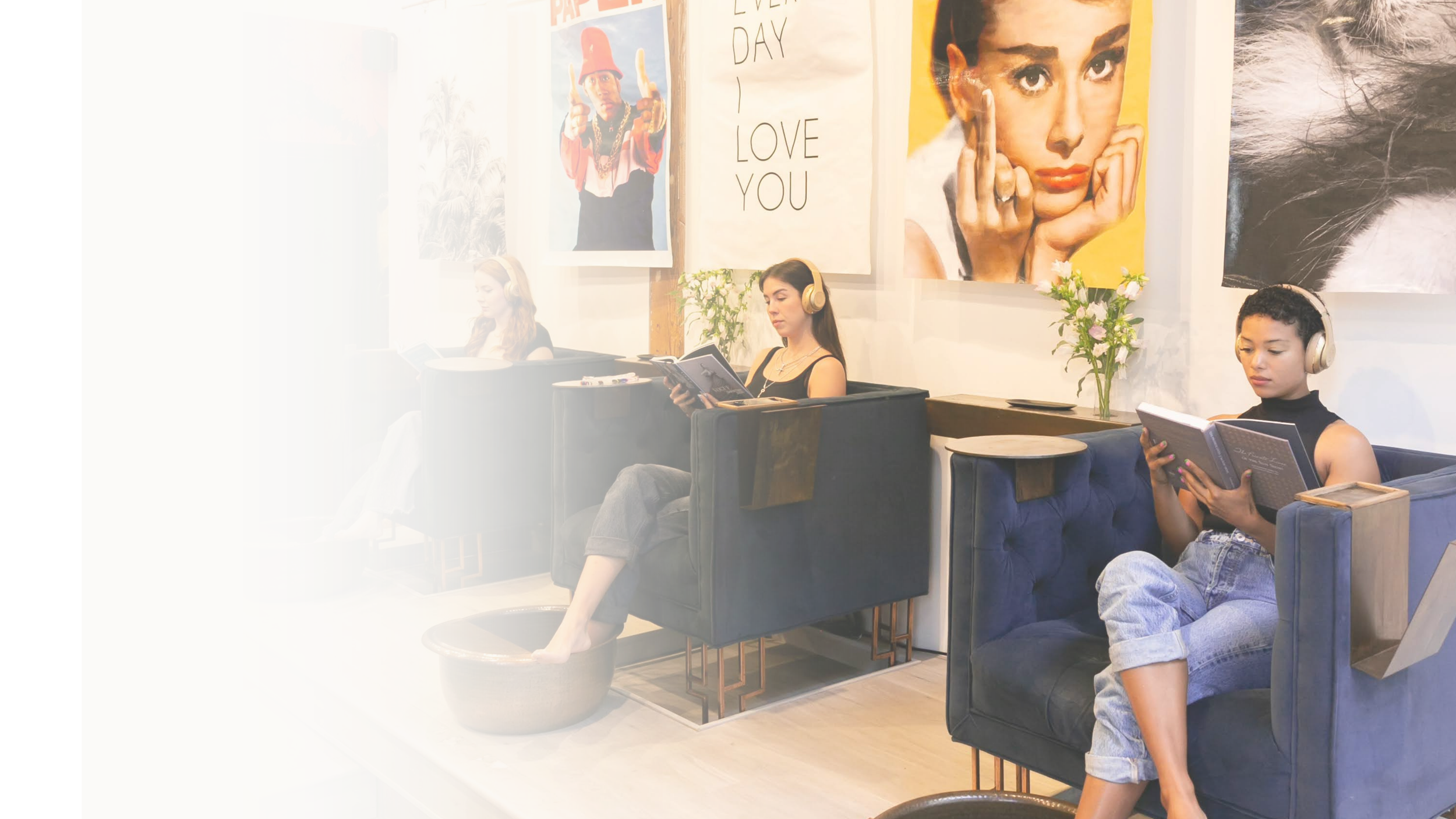 Four women sitting in a modern café, each wearing headphones and reading books or magazines. The café has large, colorful portraits and motivational posters on the wall.