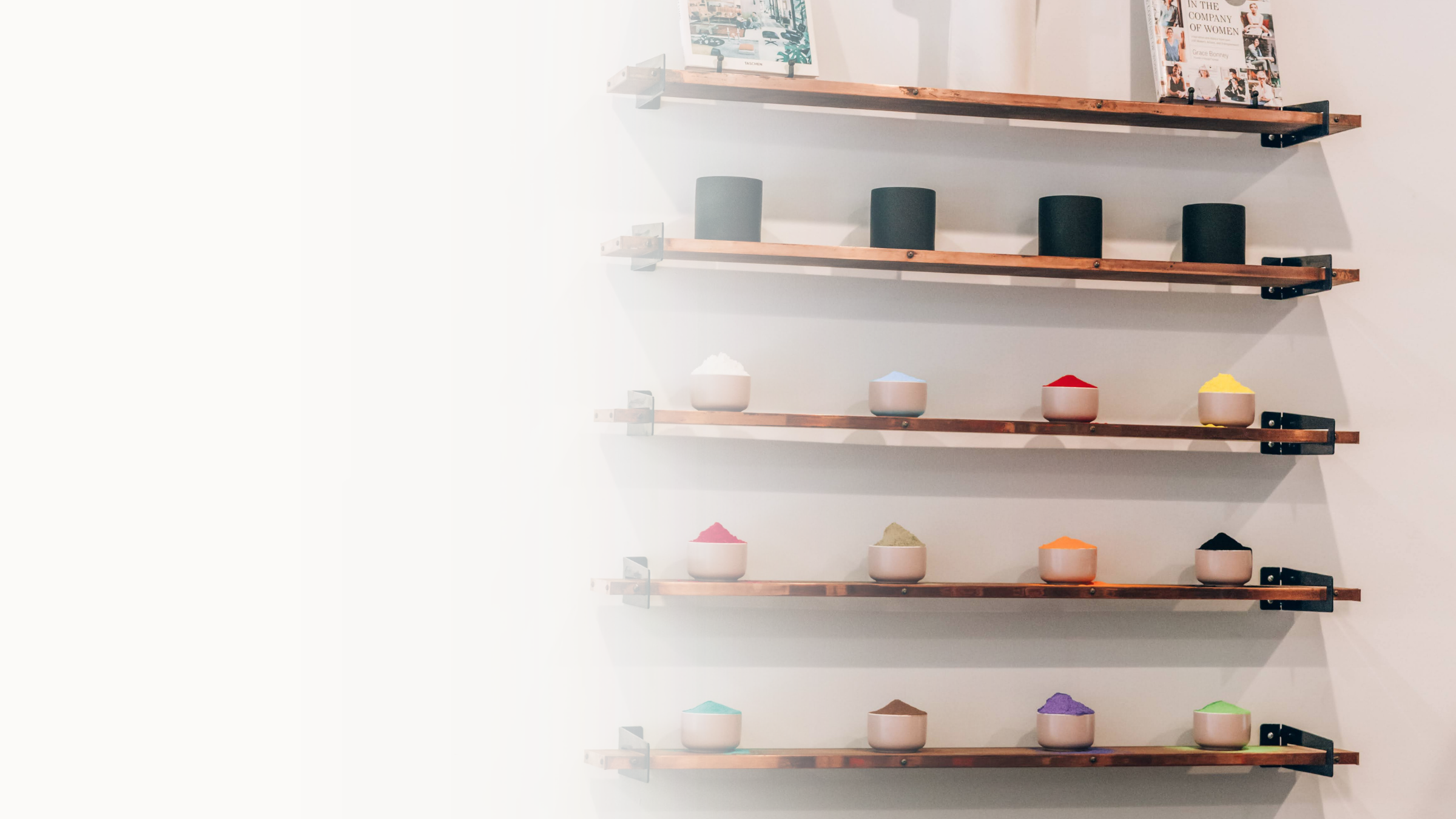 The image shows four dark-colored cups on wooden shelves mounted on a white wall. Above the shelves, there are framed pictures and magazine covers, with some shelves also holding small bowls filled with colorful powders.