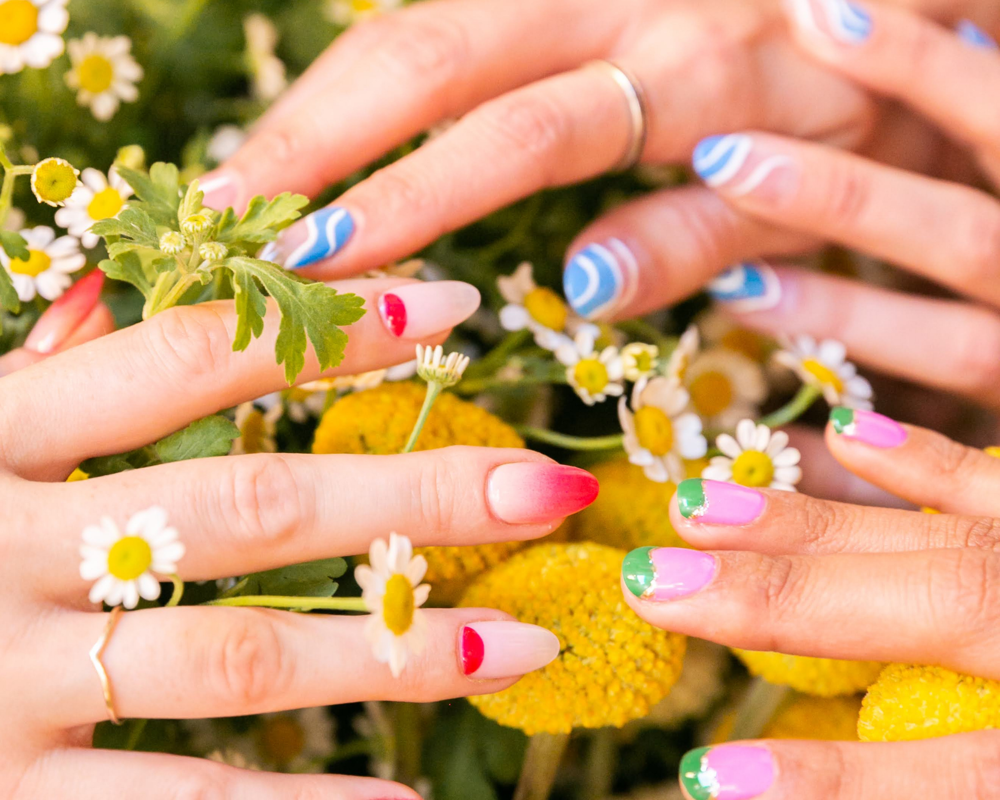 Four hands with colorful painted nails touching a bouquet of daisies and yellow flowers.