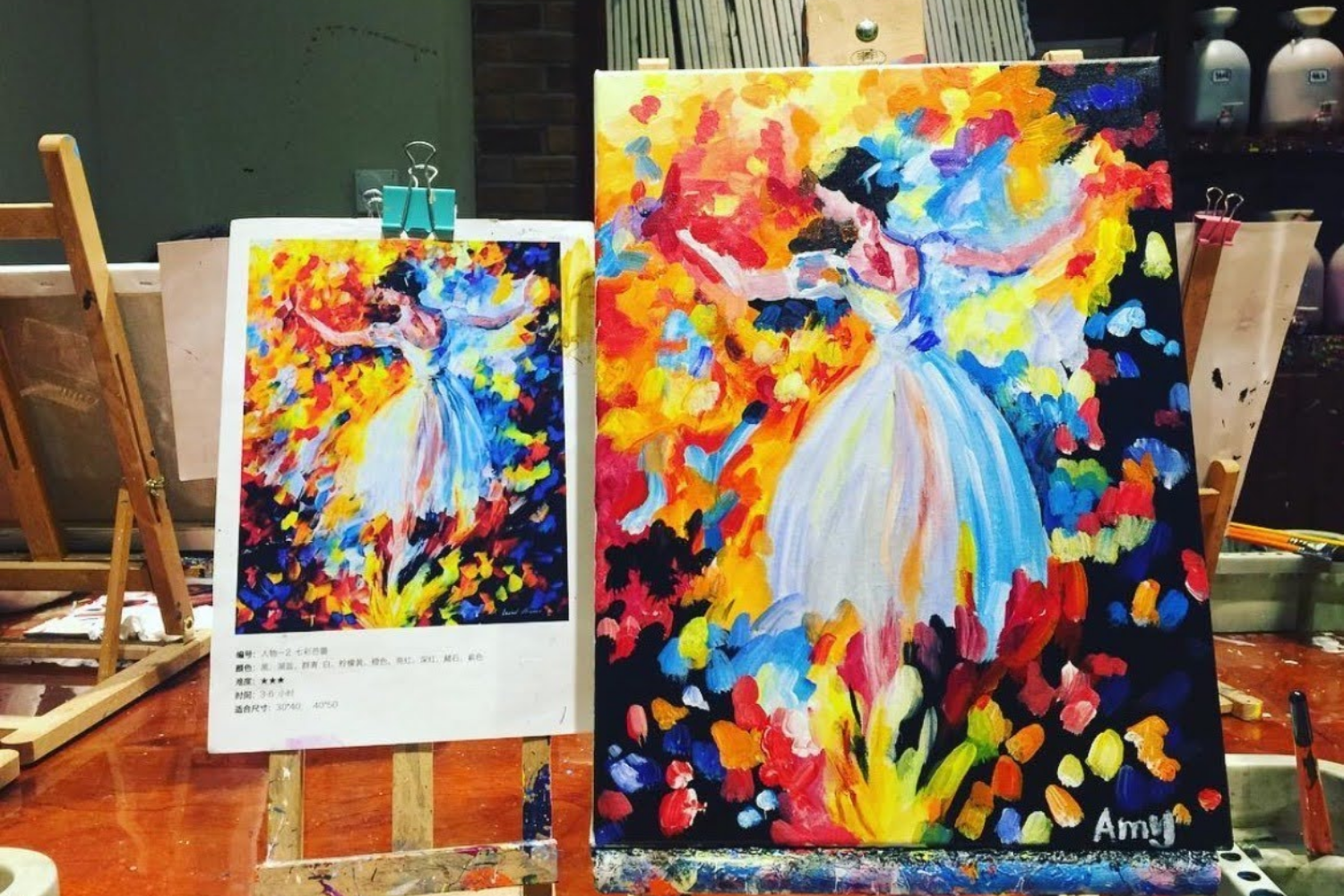 Two colorful paintings of ballerinas dancing among vibrant, abstract fall foliage. The paintings are on display, with the smaller on a white easel and the larger on a black easel. The artwork is expressive and use bold brushstrokes, signed 'Amy' on the larger painting.