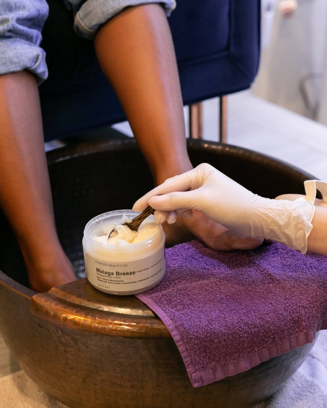 Person soaking feet in a copper foot bathtub while a technician applies lotion with a spatula.