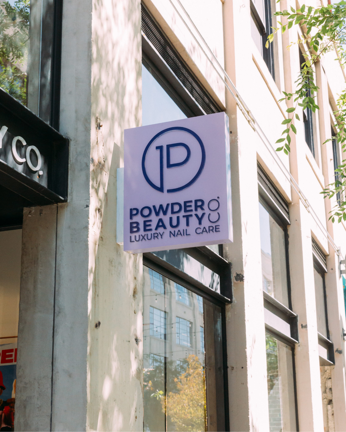 Sign for Powder Beauty, a luxury nail care salon, mounted on the exterior of a building with large windows and surrounding greenery.