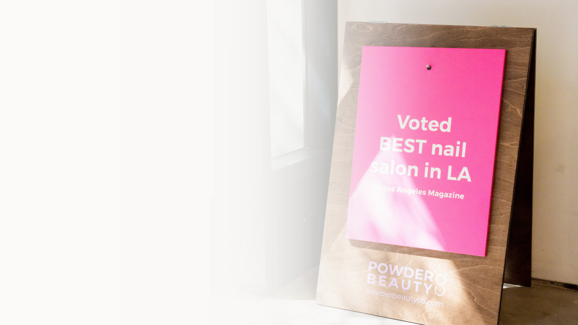 Pink signboard with white text reading 'Voted BEST nail salon in LA' on a wooden stand near a window.