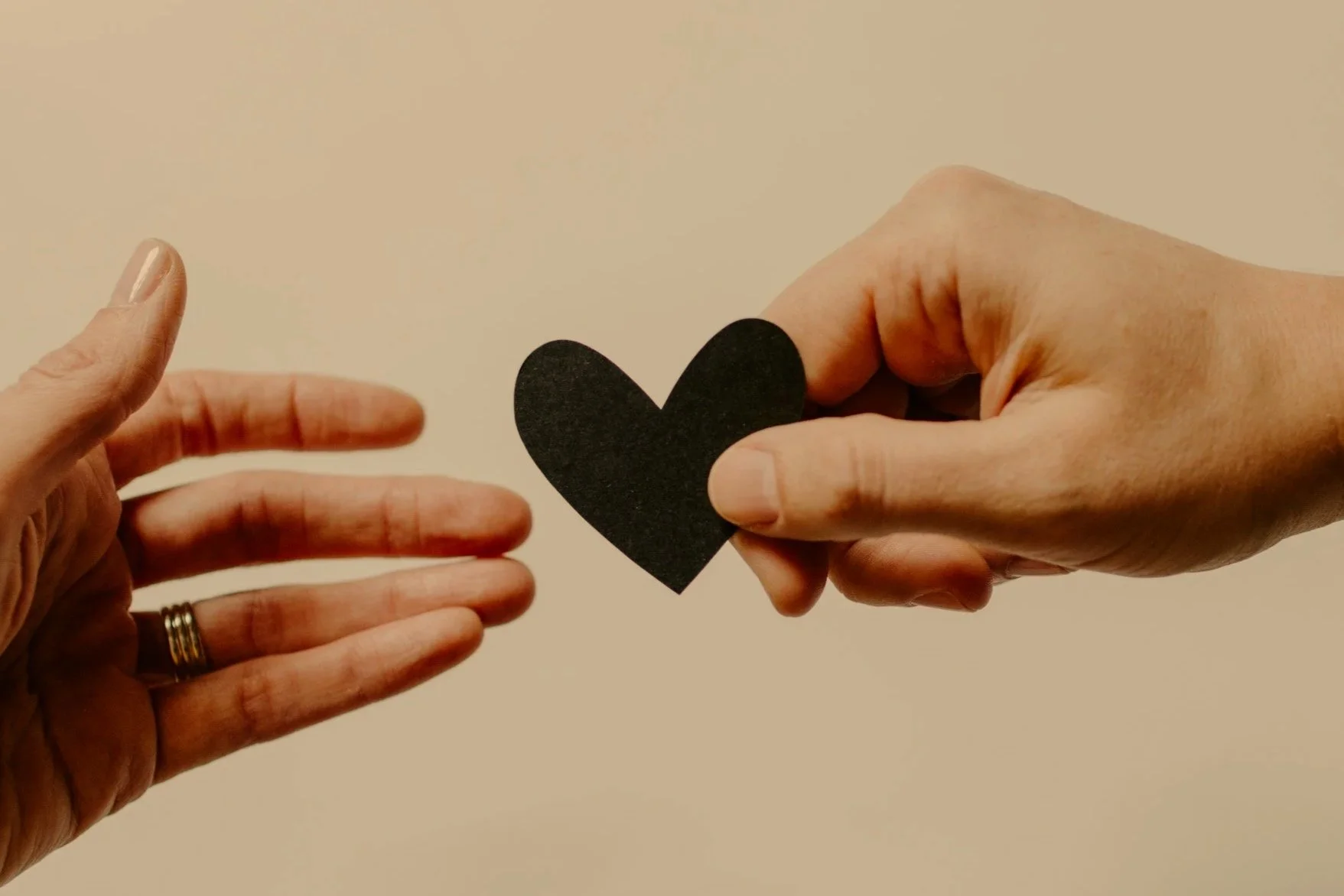 Person holding a black paper heart with their right hand, and another person extending their left hand towards it against a beige background.