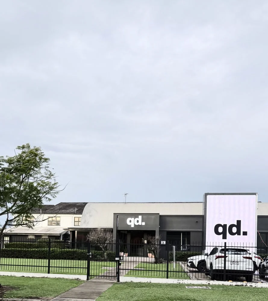 Modern building with a large sign displaying the letters 'qd.' in black, located in a parking lot with cars and a green lawn.