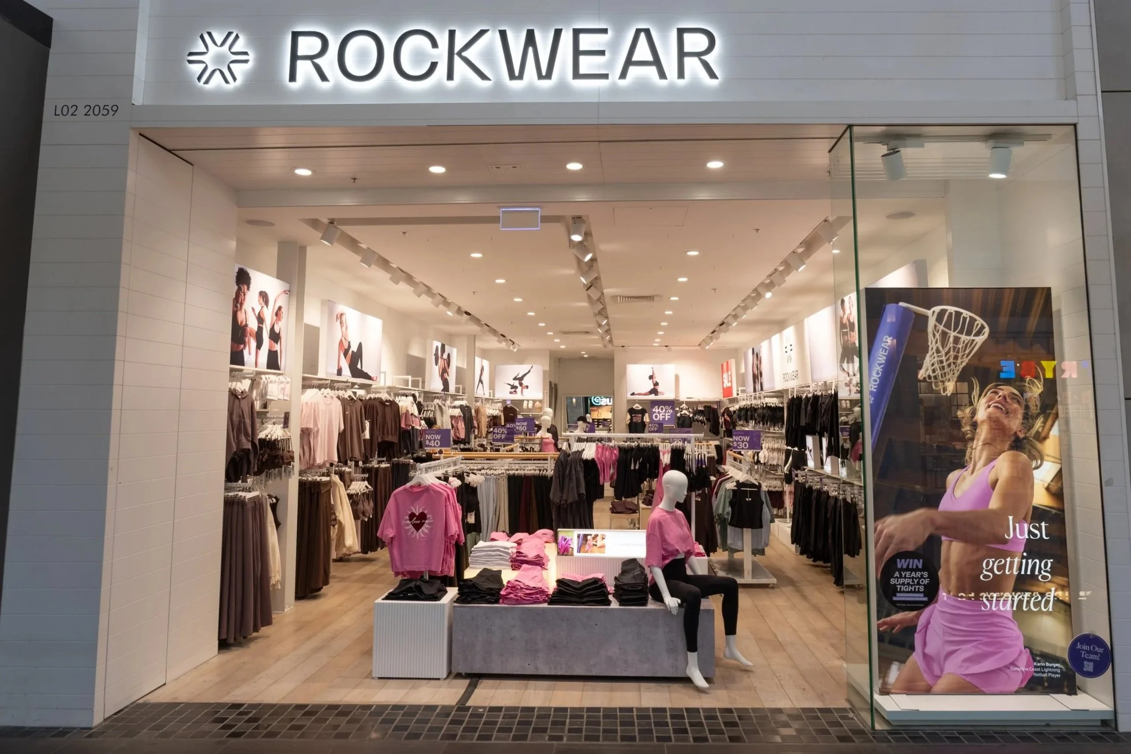 Entrance of a clothing store named ROCKWEAR with racks of clothes inside, mannequins dressed in athletic clothing, promotional signs, and a promotional poster with a happy woman in a pink top.