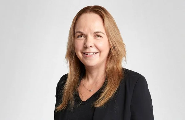 A woman with long light brown hair smiling, wearing a black top and blazer against a plain light gray background.