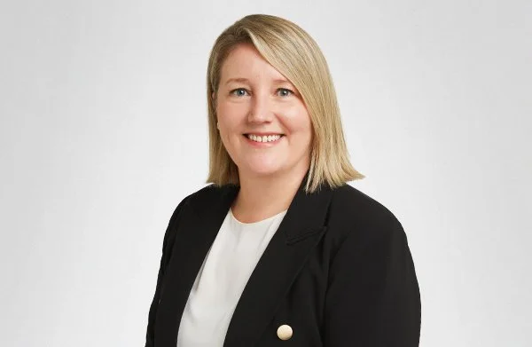 Professional portrait of a woman with blonde hair, wearing a black blazer and white blouse, smiling against a plain white background.