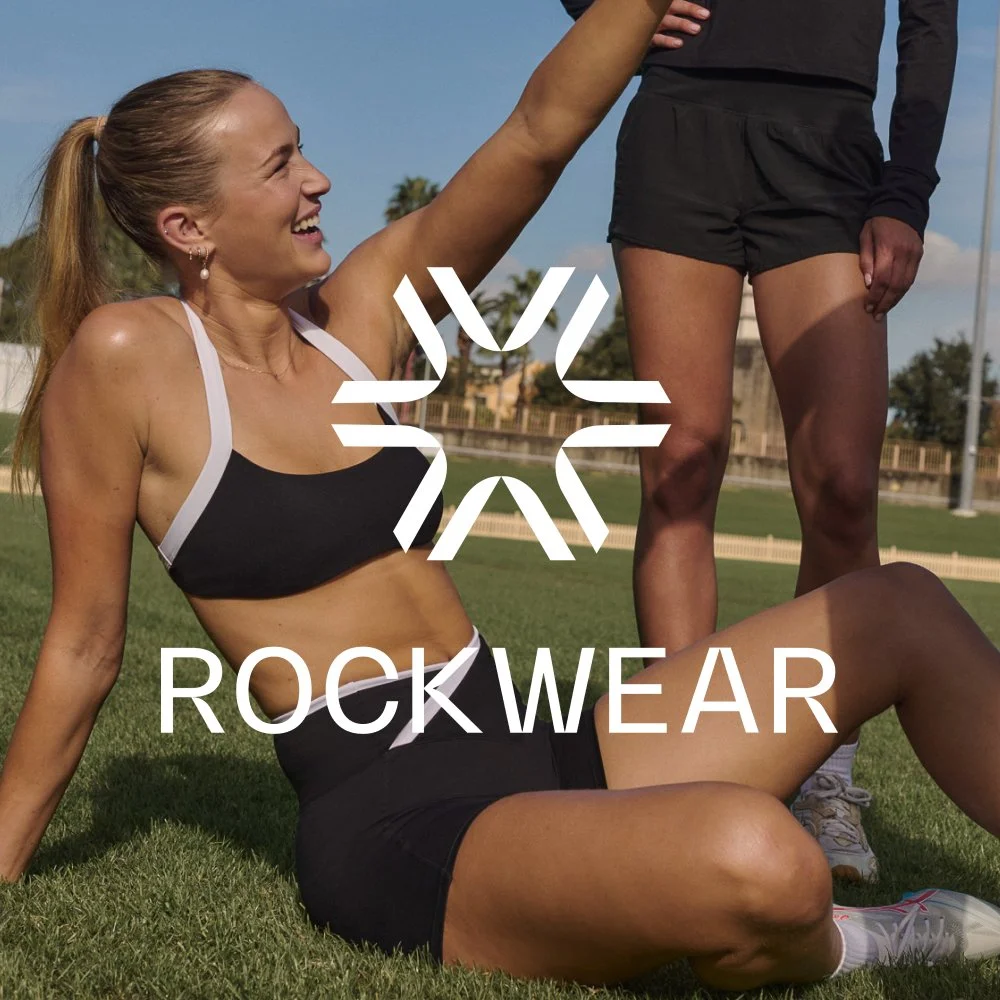 A woman in athletic wear sitting on grass, smiling and reaching out to another woman who is standing nearby, both outdoors on a sunny day. The word 'ROCKWEAR' is written across the image.